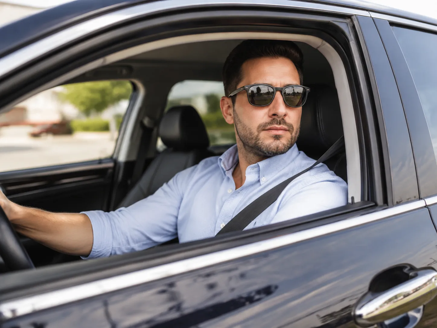 A man with a beard and sunglasses sitting in the driver's seat of a car on a sunny day, looking out the window